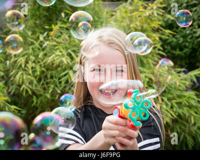 Zehn Jahre alte Blondine verwendet eine kleine Maschine zu blubbern an der Kamera in einem Garten Stockfoto