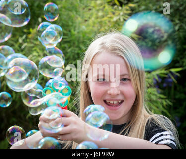 Zehn Jahre alte Blondine verwendet eine kleine Maschine zu blubbern an der Kamera in einem Garten Stockfoto