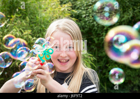 Zehn Jahre alte Blondine verwendet eine kleine Maschine zu blubbern an der Kamera in einem Garten Stockfoto