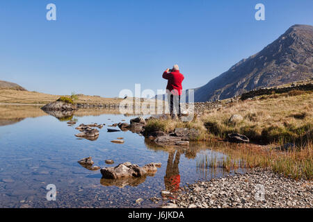 Klar, Senior woman in fotografieren auf sein Handy an Llyn Idwal, Snowdonia, North Wales, UK, an einem schönen sonnigen Frühlingstag mit roten Fleece-Jacke Stockfoto