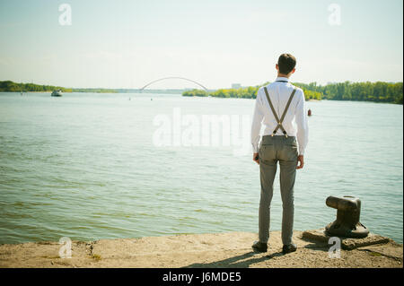 junge magerer Mann, elegant stilvoll gekleidet in weißem Hemd, graue Hose mit Hosenträgern und Fliege. Wegsehen, an der Brücke am Wasser und s Stockfoto