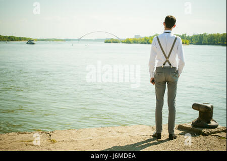 junge magerer Mann, elegant stilvoll gekleidet in weißem Hemd, graue Hose mit Hosenträgern und Fliege. Wegsehen, an der Brücke am Wasser und s Stockfoto