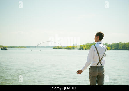 junge magerer Mann, elegant stilvoll gekleidet in weißem Hemd, graue Hose mit Hosenträgern und Fliege. Wegsehen, an der Brücke am Wasser und s Stockfoto