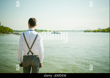 junge magerer Mann, elegant stilvoll gekleidet in weißem Hemd, graue Hose mit Hosenträgern und Fliege. Wegsehen, an der Brücke am Wasser und s Stockfoto