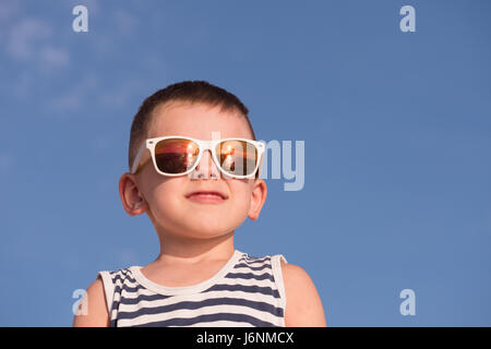 lächelnde Knabe tragen weiße Sonnenbrille mit Meer Sonnenuntergang Reflexion und Sailor Streifen auf blauem Himmelshintergrund mit Exemplar Weste Stockfoto