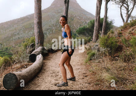 Voller Länge Schuss der glückliche junge Asiatin steht auf Bergweg und lächelnd. Fitness-Frau in Sportkleidung auf Kreuz Feldweg. Stockfoto