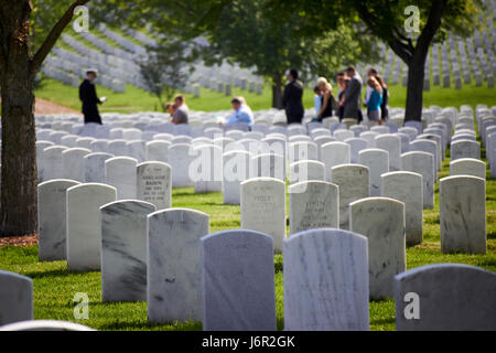 Reihen von weißen Grabsteinen unter Bäumen mit Trauerfeier im Hintergrund auf dem Arlington Cemetery Washington DC USA Stockfoto