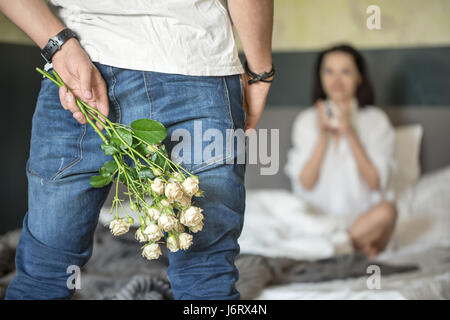 Mann in Blue Jeans und weißes T-shirt hält einen Strauß Rosen hinter ihrem Rücken. Er befindet sich gegenüber seinem Mädchen in einem weißen Hemd, das auf dem Bett sitzt. Closeu Stockfoto