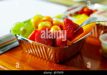 Gesunder Mensch mit einer Gabel um zu Essen im Büro einen köstlichen frischen Obstsalat auf Aluminiumbox auf Holztisch Stockfoto
