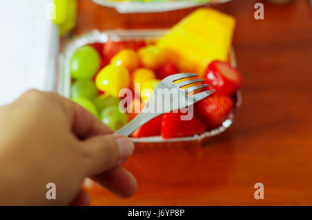 Gesunder Mensch mit einer Gabel um zu Essen im Büro einen köstlichen frischen Obstsalat auf Aluminiumbox auf Holztisch über Ansicht Stockfoto