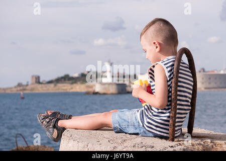 Schöne kleine Junge in einem Seemann Streifen Weste und shorts auf konkrete Wellenbrecher vor dem Hintergrund der Seehafen Standortwahl und Fernglas in seinem Besitz Stockfoto