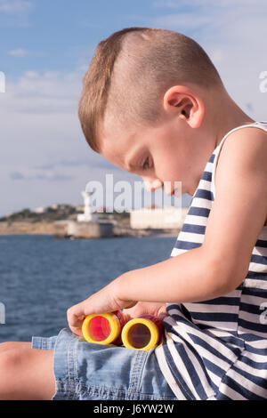 niedliche kleine Junge in Seemann Singulett Spiel mit bunten Fernglas auf dem Hintergrund des Meeres und der Leuchtturm Stockfoto