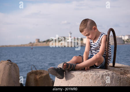 Schöne kleine Junge in Sailor Streifen Weste entspannend sitzen gegen das Meer Hafen mit Leuchtturm Stockfoto