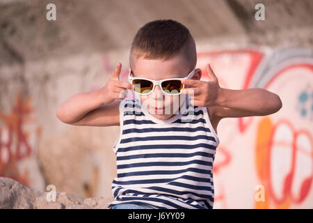 Lustige kleine Junge in einer Sonnenbrille und Streifen Matrosenhemd auf Graffiti wall Hintergrund Stockfoto