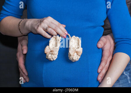 Nahaufnahme von jungen Paar weiße schwangere Frau im blauen Kleid mit Baby Babyschuhe auf Bauch und Mann die Hände an den Hüften. Stockfoto