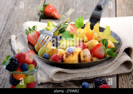 Köstliche österreichische "Kaiserschmarren" (wie zerkleinerte süße Pfannkuchen) serviert mit frischen bunten Früchten in einer eisernen Pfanne auf Vintage Leinen Stockfoto