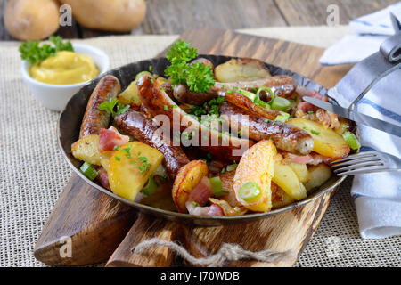 Gebratene Nürnberger Würstchen mit knusprigen Bratkartoffeln und Speck, serviert in einer eisernen Pfanne auf einem Holzbrett Stockfoto