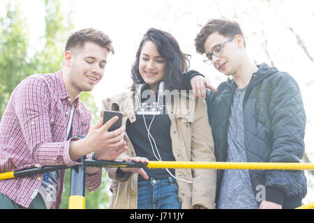 Drei junge Menschen lächelnd zu sprechen und haben viel Spaß. Anzeigen von Bildern auf R-Handy. Linseneffekt. Stockfoto
