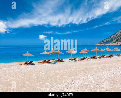 Sommer-morgen-Strand mit türkisblauem Wasser und bewölkten Himmel, Sonnenliegen und strohig Sonnenschirme (Albanien). Stockfoto