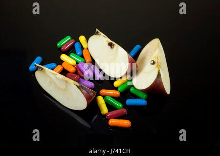Scheiben von rotem Apfel, umgeben von bunten Pillen oder Drogen, Konzeptbild über Ernährung und gentechnisch veränderten Lebensmitteln auf schwarzem Hintergrund Stockfoto