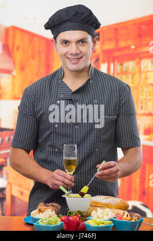 Gut aussehend lächelnder Mann hält in einer hand eine köstliche Birne bedeckt mit Schokoladen-Fondue und in der anderen Hand eine Tasse Weißwein mit verschiedene Früchte wie Erdbeere, Birne, Kiwi, Apfel und Brotscheiben in Schalen auf Holztisch Stockfoto