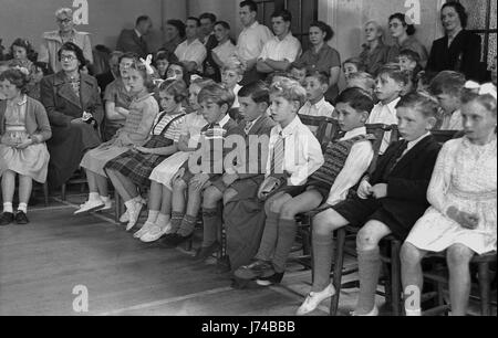 In den 1950er Jahren, histroisch, ein Publikum von Grundschulkindern, Lehrern und Eltern beobachten eine Musik- und Bewegungsvorstellung in der Schulhalle, England, Großbritannien. Einige der Kinder sind in Tneir-Uniform, während ihre Füße, einige tragen Sandalen und Pilmolls. Stockfoto