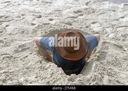 Ein junger Mann versucht, sich aus einem Loch in den Sand zu graben, die er sich an einem Strand eines sonnigen Tages bekommen hat Stockfoto