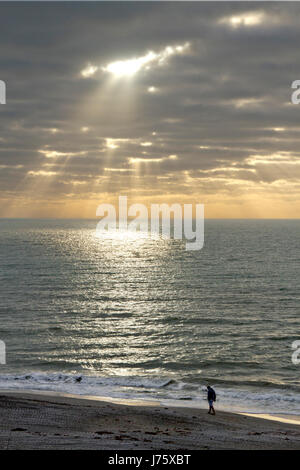 Lichtstrahlen unverdünnt in durch dunkle Wolken auf einem ruhigen Meer Iluminating einen leuchtenden Kreis des Lichts zu brechen, wie ein einsamer Mann ruhig Strand geht Stockfoto