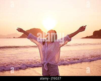 junge asiatische Frau, die am frühen Morgen Sonnenlicht mit offenen Armen am Strand zu genießen. Stockfoto