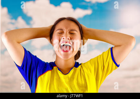 Jungen ecuadorianischen Frau mit offiziellen Marathon Fußball Shirt stehen vor der Kamera, sehr engagierte Körpersprache beobachten Spiel mit großer Begeisterung, blauer Himmel und Wolken-Hintergrund Stockfoto