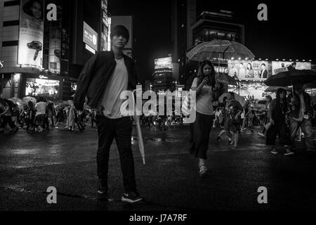 Schwarz / weiß Nacht Foto von Fußgänger auf der belebten Shibuya Crossing. Ein Mädchen, SMS und tragen einen Regenschirm, leicht hinter ein junger Mann geht. Stockfoto