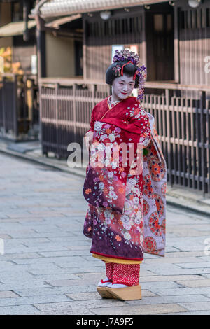 Geisha in einer farbenfrohen Kimono, weiße Make-up auf ihr Gesicht & Holzsandalen auf ihre Füße angehoben. Auf dem Kopf, einen wunderschönen Haarschmuck.  Outdoor-Bild Stockfoto