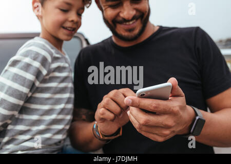 Nahaufnahme der glückliche Vater und Sohn vor dem Auto auf der Suche auf dem Handy. Junger Mann und kleiner Junge mit Smartphone während auf Roadtrip. Stockfoto