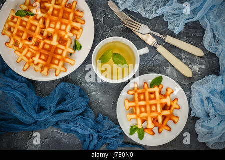 Zwei Platten mit belgischen Waffeln garniert mit Minze, Glas Tasse grüner Tee mit Minze, Gabel mit Messer auf grauem Hintergrund. Ansicht von oben Stockfoto