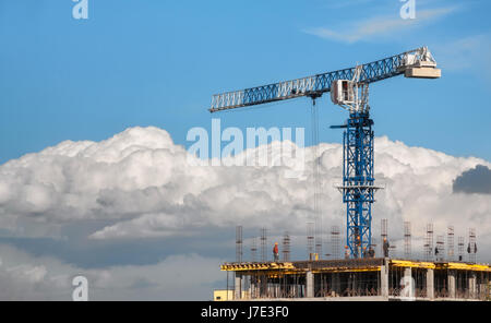 Die Bau-Turmdrehkran ist blau neben den Bauherren auf dem Dach eines Gebäudes im Bau. Auf einem Hintergrund von Wolken und blauer Himmel Stockfoto