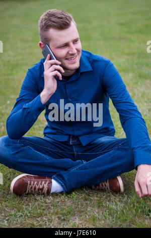 Junger Mann mit blonden Haaren, auf dem Rasen und Gespräche über ein Handy, ein Lächeln auf seinem Gesicht sitzen Stockfoto