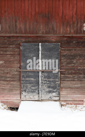 Nahaufnahme alter verwitterter hölzerner Scheunentüren gegen rote Holzwände im Winter, die rustikale Textur und skandinavisches Erbe zeigen Stockfoto
