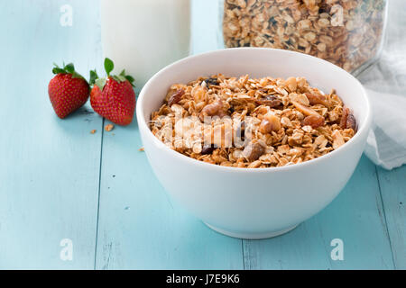 Hausgemachte gebackene Müsli in eine Schüssel geben und in ein Glas, eine Flasche Milch und Erdbeeren auf einem türkisfarbenen Holztisch. Gesundes Frühstück Stockfoto