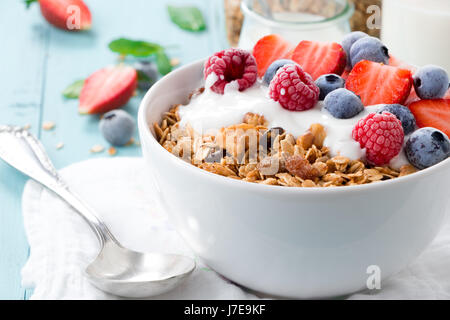 Gesundes Frühstück in einer Schüssel mit hausgemachtem gebackenes Müsli, gefrorene Beeren, frische Erdbeeren und Joghurt auf einem türkisfarbenen Holztisch Stockfoto