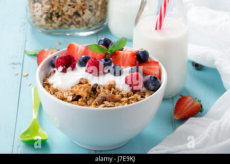 Gesundes Frühstück in einer Schüssel mit hausgemachtem gebackenes Müsli, gefrorene Beeren, frische Erdbeeren und Joghurt auf einem türkisfarbenen Holztisch Stockfoto