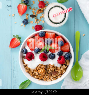 Gesundes Frühstück in einer Schüssel mit hausgemachtem gebackenes Müsli, gefrorene Beeren, frische Erdbeeren, eine Flasche Milch und Joghurt auf einem türkisfarbenen Holztisch Stockfoto