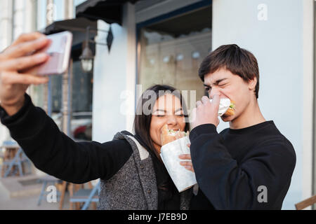 Bruder und Schwester machen Selfie mit geschlossenen Augen während des Essens Burger auf der Straße Stockfoto