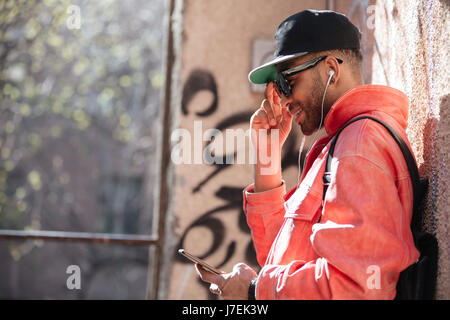 Porträt eines schönen Hipster Afro amerikanische Sonnenbrillen hören Musik mit dem Handy und Kopfhörer während stützte sich auf einer Straße Wand Stockfoto