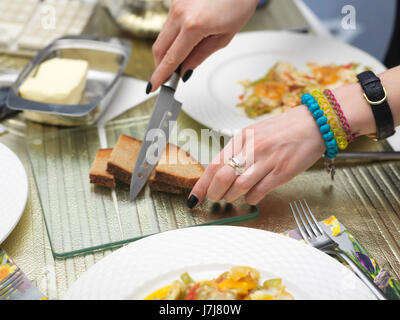 Weibliche Hände ausbreitende Butter aufs Brot, einen unordentlichen Frühstückstisch als Hintergrund. Stockfoto
