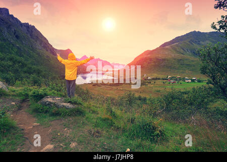 Panorama Blick auf den Fjord. Twilight-Zeit mit einer erstaunlichen orangefarbenen Himmel. Der junge Mann mit den Händen in der Luft stehend auf einer Klippe aus Rock und blicken Sonnen Stockfoto