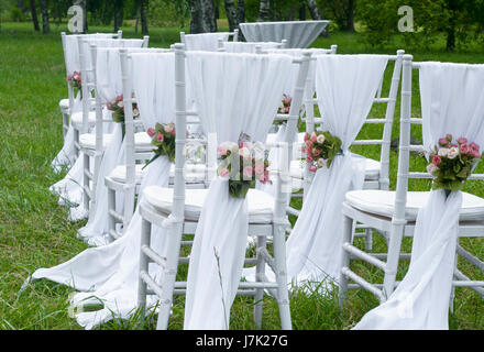 Weiße Stühle stehen mit Blumen im Park in den Zeilen vor dem Traualtar. Stockfoto