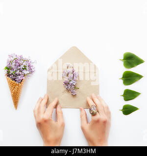 Quadratische Karte mit Frauen Hände berühren Handwerk Papierumschlag mit lila Blüten, umgeben von einer Waffel Mais mit einem Bouquet von Flieder und einer Reihe von Grün l Stockfoto
