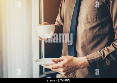 Junger Geschäftsmann hält Tasse Kaffee im Büro am Fenster. Stockfoto