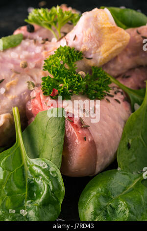 Vertikale Foto von rohem Hühnerfleisch Beine. Drei Stücke von Fleisch sind auf dunkle Vintage Tablett gelegt. Grüne Kräuter wie Petersilie und Spinat sind um mit verschüttet. Stockfoto