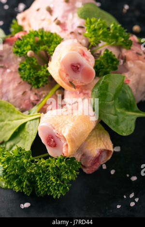 Vertikale Foto von rohem Hühnerfleisch Beine. Drei Stücke von Fleisch sind auf dunkle Vintage Tablett gelegt. Grüne Kräuter wie Petersilie und Spinat sind um mit verschüttet. Stockfoto